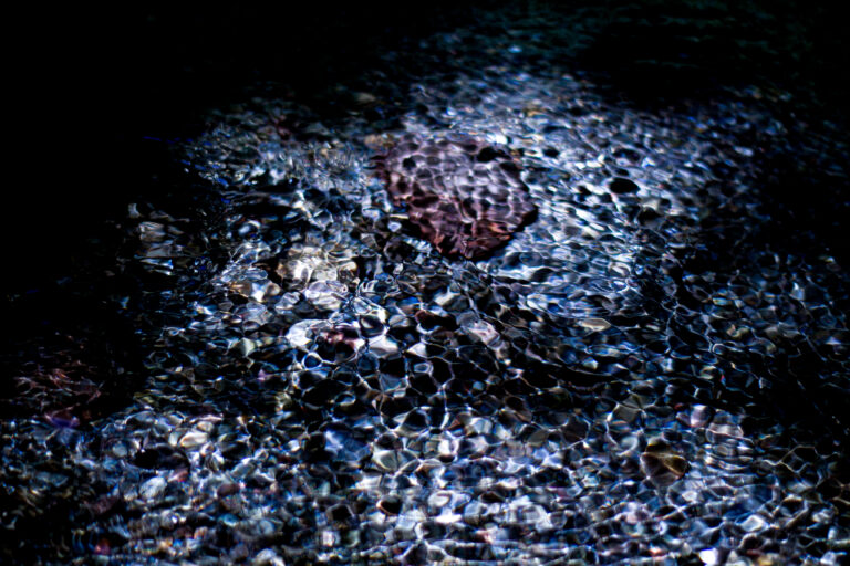 Fine art photograph of a shallow river where rippling reflections transform stones into shimmering, organic patterns beneath a dark surface. The scene evokes a quiet, dreamlike atmosphere between abstraction and reality.