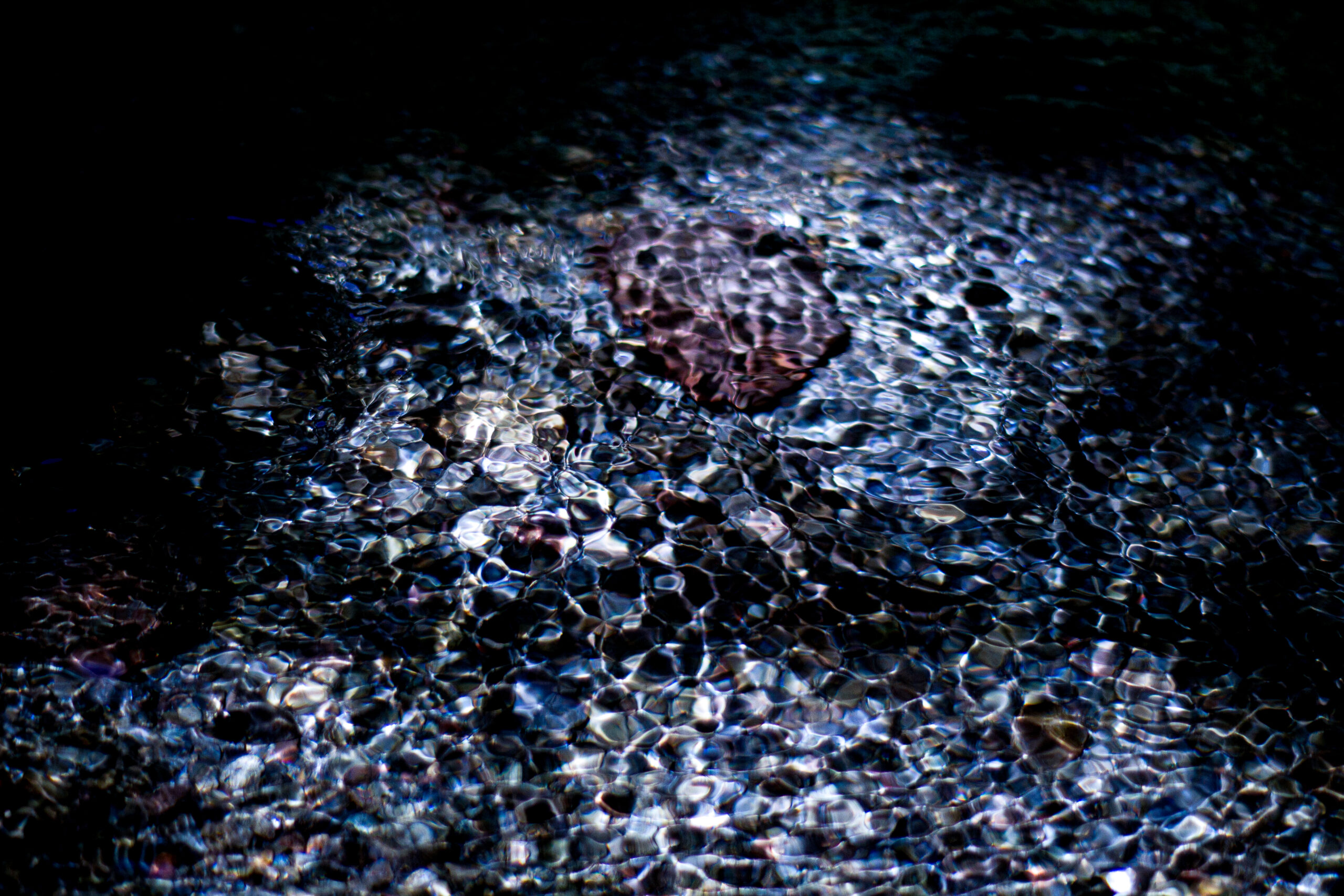 Fine art photograph of a shallow river where rippling reflections transform stones into shimmering, organic patterns beneath a dark surface. The scene evokes a quiet, dreamlike atmosphere between abstraction and reality.