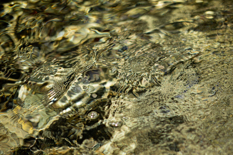 Abstract photograph of a shallow stream where soft golden light, gentle ripples, and a few small air bubbles blur the boundary between reflection and the riverbed, creating a calm, dreamlike surface.