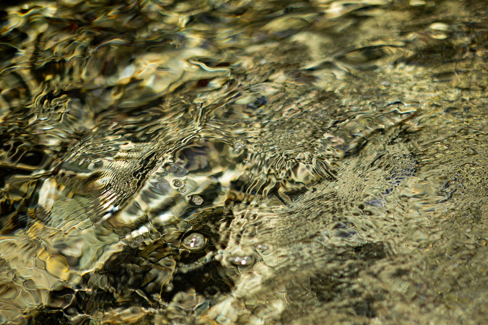 Abstract photograph of a shallow stream where soft golden light, gentle ripples, and a few small air bubbles blur the boundary between reflection and the riverbed, creating a calm, dreamlike surface.