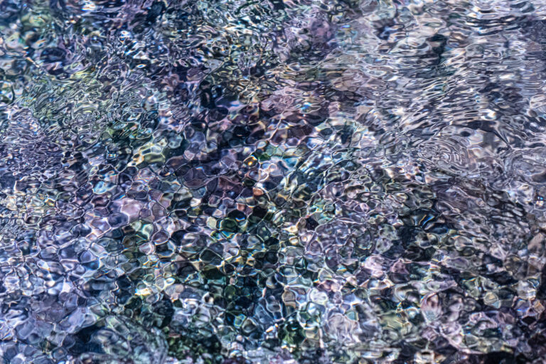 Abstract fine art photograph of a shallow stream where rippling water transforms stones and reflections into translucent, cell-like patterns in muted blue, green, and violet tones.