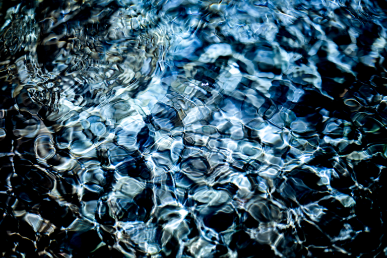 Close-up abstract photograph of sunlight fracturing across rippling blue water, creating a fragmented, glass-like pattern of light and shadow