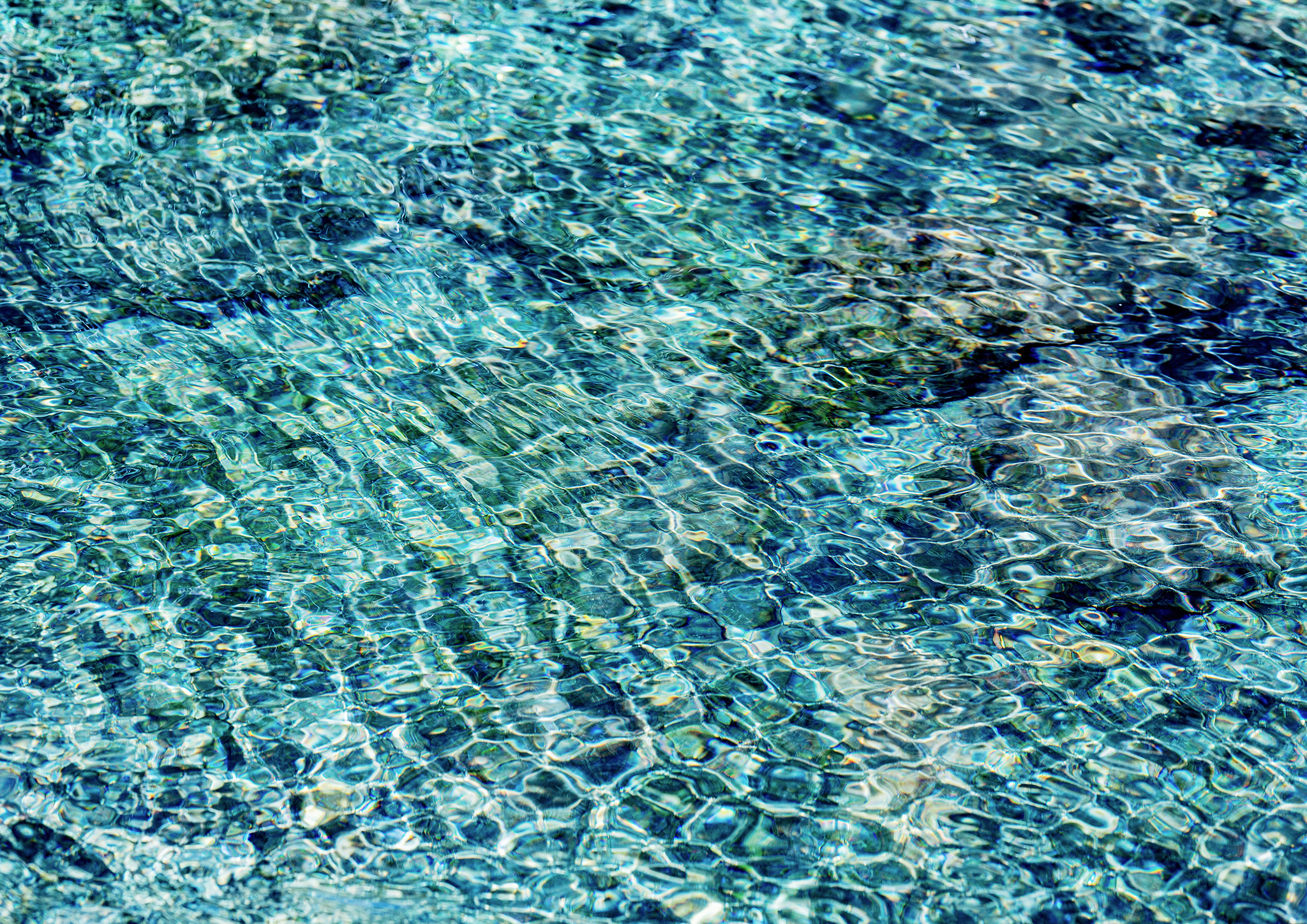 Clear blue river water with shimmering sunlight over submerged stones, creating abstract patterns of light and gentle ripples.