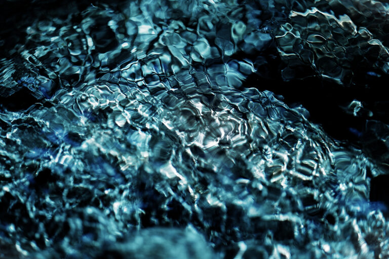 A close-up abstract photograph of flowing river water, where deep blue and black tones merge with shimmering reflections, creating organic patterns that resemble glass, stone, and living cells.