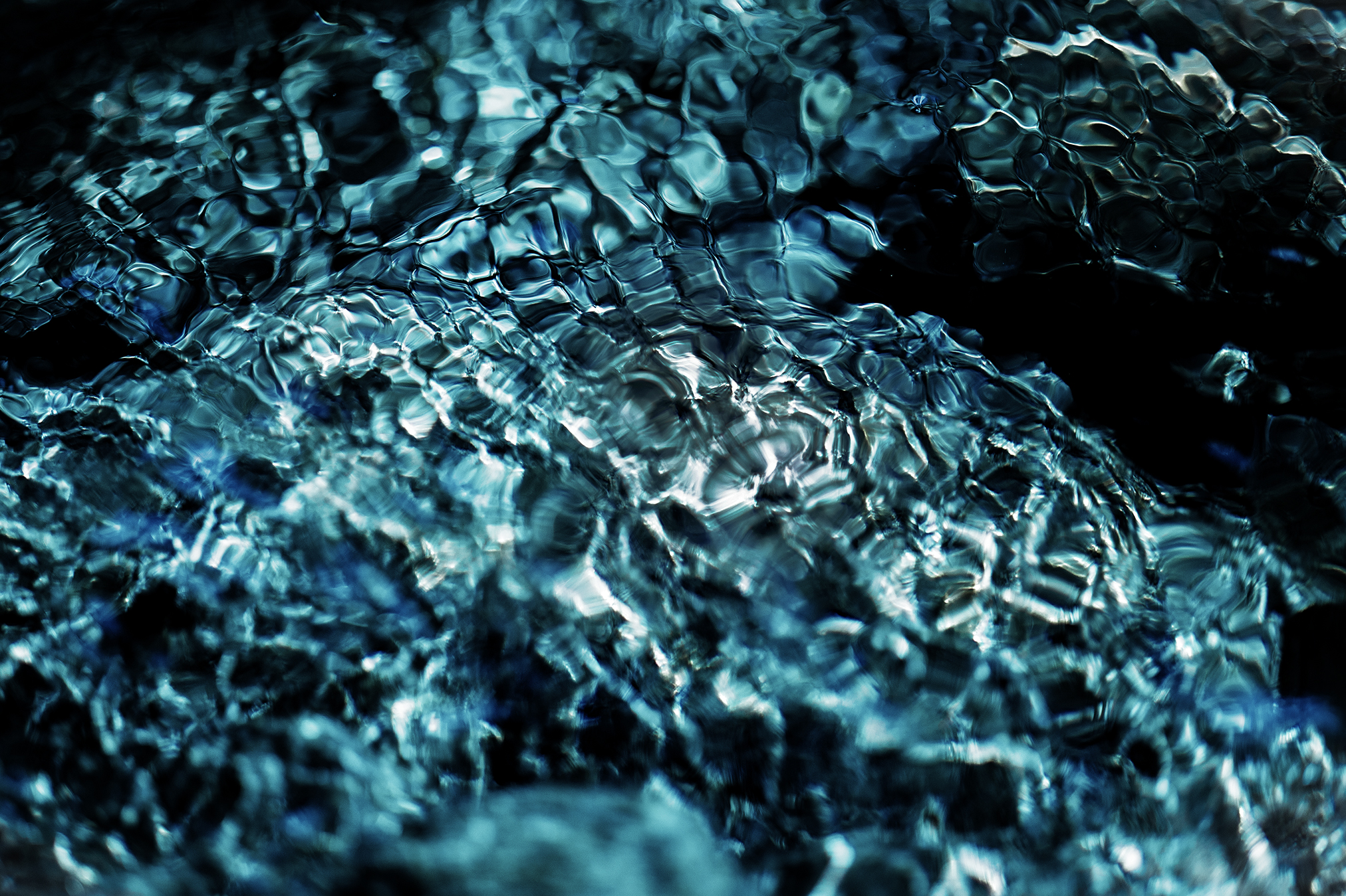 A close-up abstract photograph of flowing river water, where deep blue and black tones merge with shimmering reflections, creating organic patterns that resemble glass, stone, and living cells.